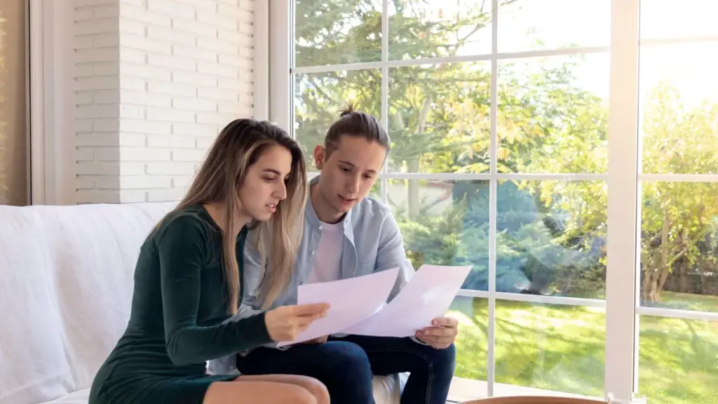 Casal jovem sentado no sofá lê com atenção as folhas de um contrato, perto de uma janela ampla