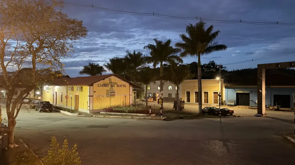 Estação ferroviária de Carmo da Mata, prédio amarelo com o nome da cidade, cercado por palmeiras, ao entardecer